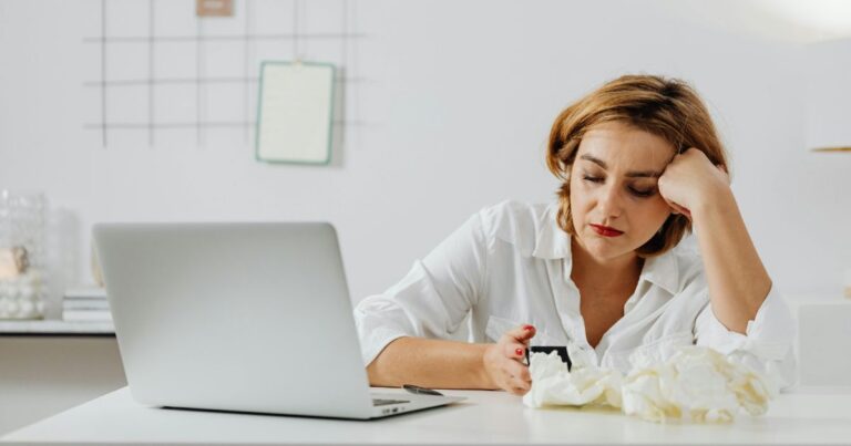 Woman feeling unmotivated and overwhelmed while sitting alone at home with her laptop and phone — representing the emotional weight of living alone and feeling unproductive.